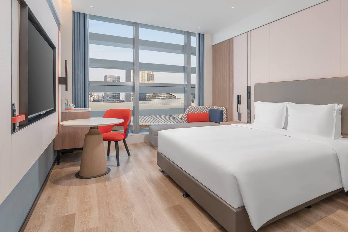 Ein Hotelzimmer mit Bett, Schreibtisch, Stuhl und einem großen Fenster mit Blick auf die Stadt.
