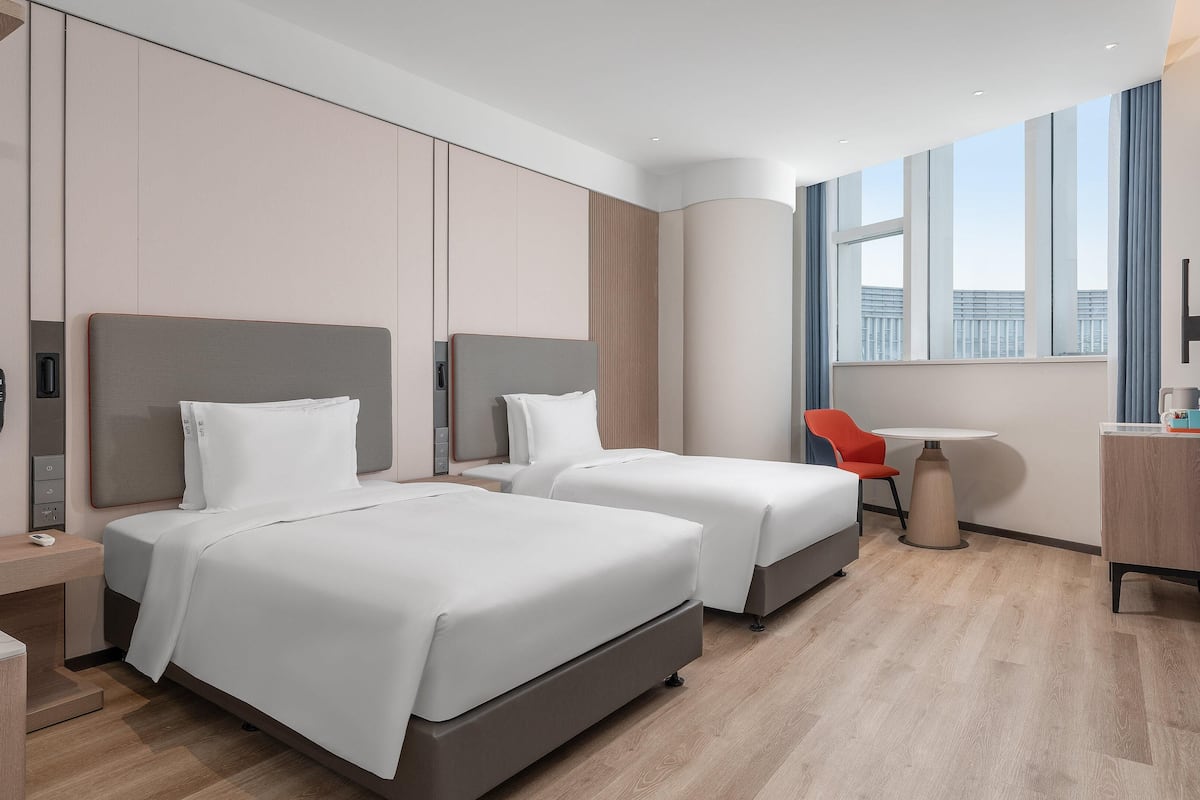 Ein modernes Hotelzimmer mit zwei Betten, einem Schreibtisch, einem Stuhl und einem großen Fenster mit Blick auf die Stadt.