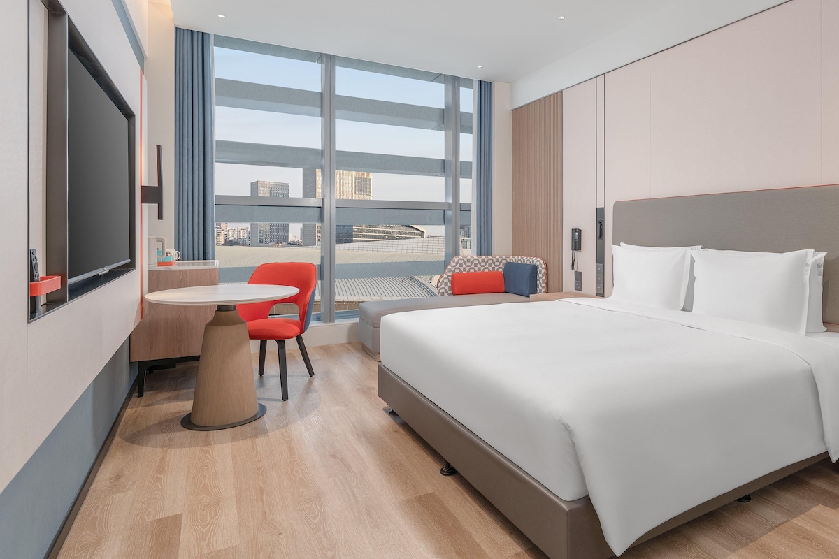 A hotel room with a bed, a desk, a chair, and a large window offering a city view.