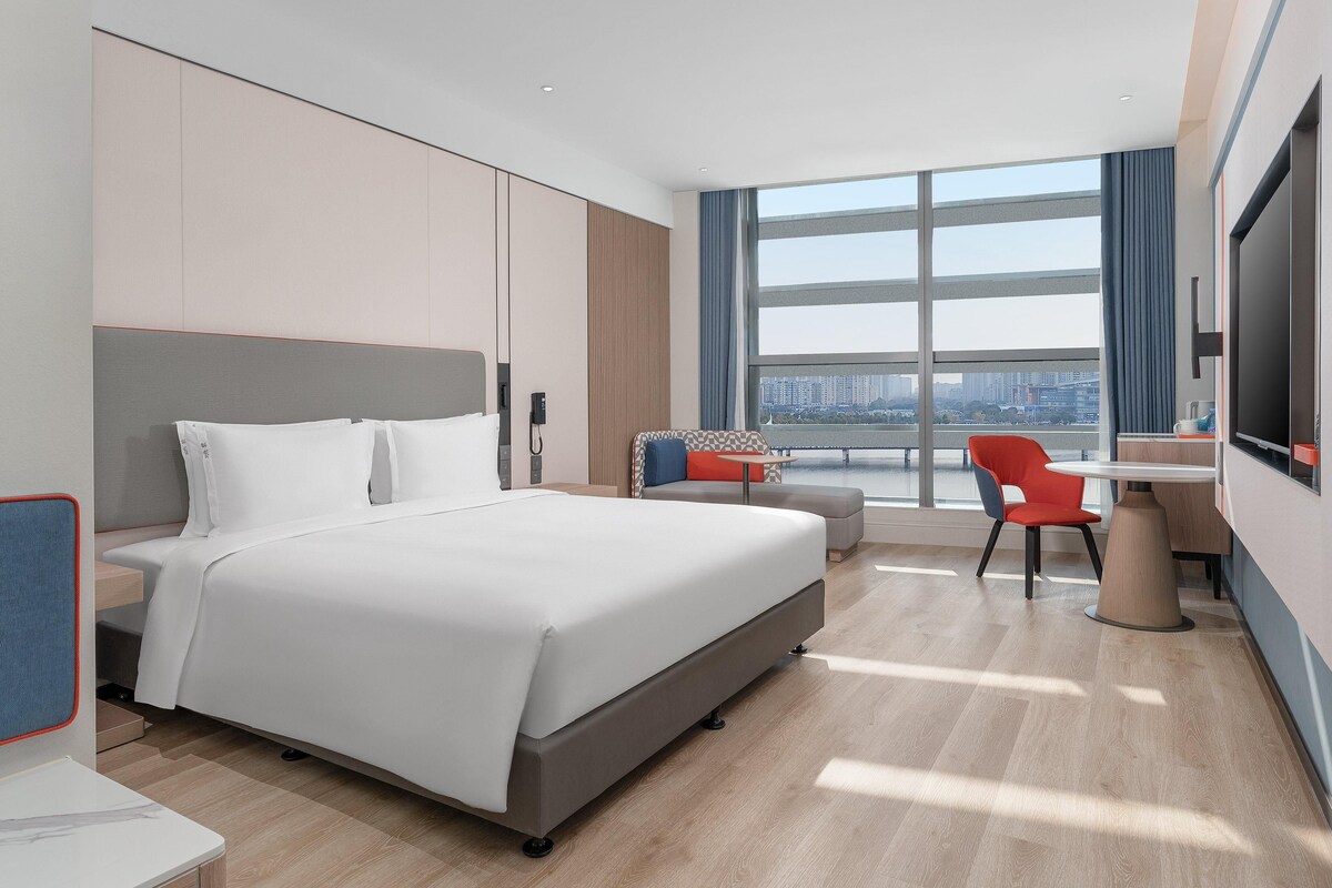 A modern hotel room with a large bed, a desk, and a chair. There is a large window with a city view.