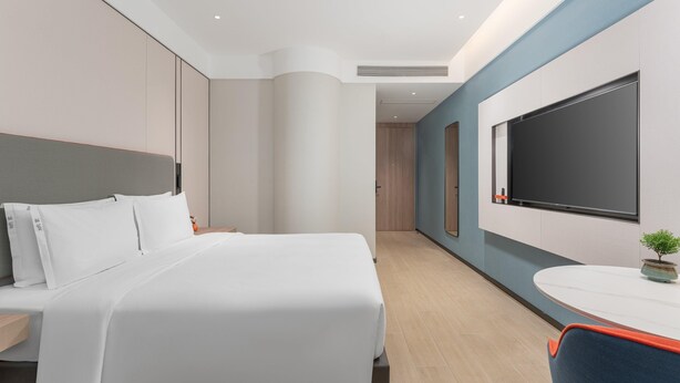 Una habitación de hotel moderna con una cama grande, un escritorio con silla, un televisor de pantalla plana montado en la pared y una pequeña planta en maceta.