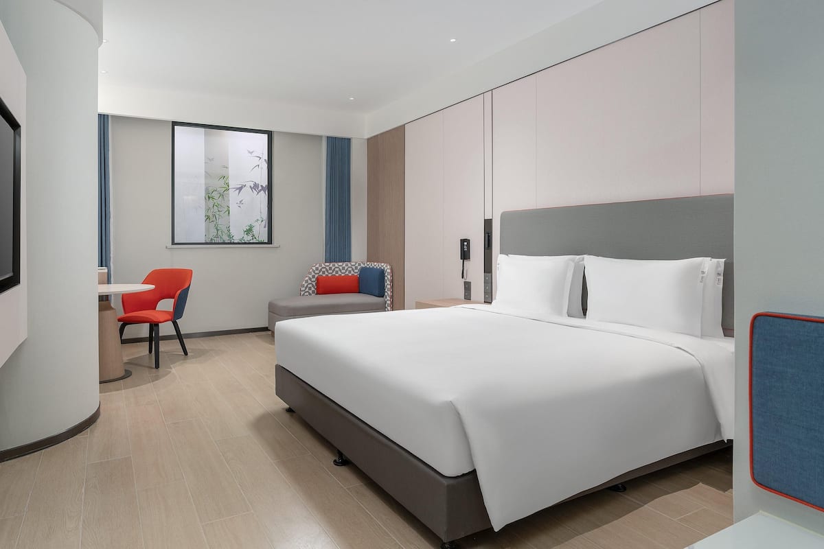 Ein modernes Hotelzimmer mit einem großen Bett, einem Schreibtisch mit Stuhl, einem Fernseher und einem Fenster mit Blick auf Bäume.
