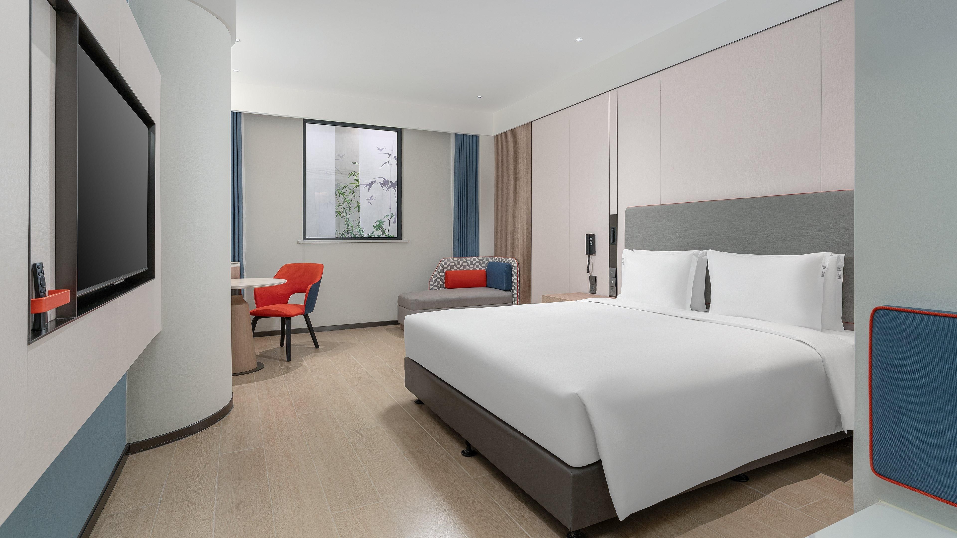 Une chambre d’hôtel moderne avec un grand lit, un bureau avec une chaise, une télévision et une fenêtre donnant sur des arbres.