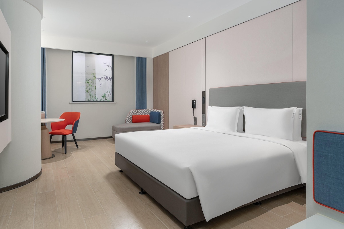 A modern hotel room with a large bed, a desk with a chair, a television, and a window with a view of trees.