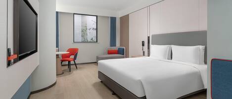 Une chambre d’hôtel moderne avec un grand lit, un bureau avec une chaise, une télévision et une fenêtre donnant sur des arbres.