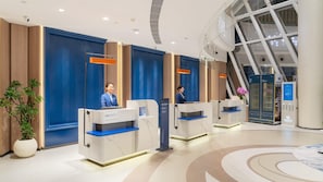 A modern hotel reception area with three reception desks, a large potted plant, and a glass elevator.