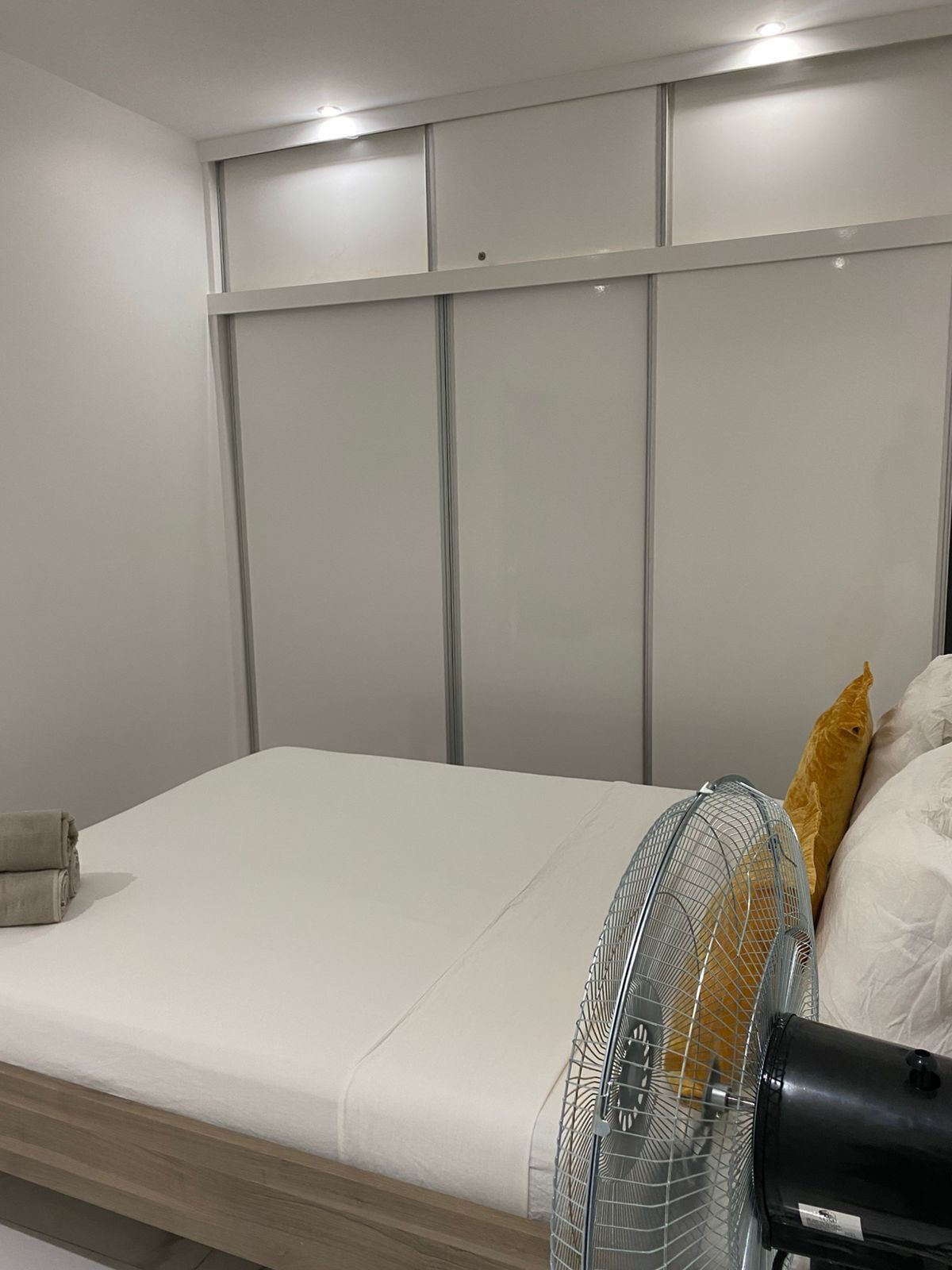 Un lit avec un matelas blanc, un ventilateur gris et une tête de lit en bois.