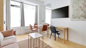 A modern living room with a sofa, a dining table with chairs, a TV mounted on the wall, and large windows with curtains.