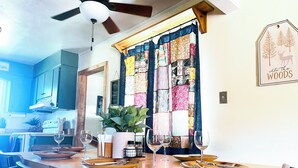 A dining area with a wooden table set for four, a potted plant, and a sign reading "Into the Woods". - Charming 3-bedroom cabin in with AC, spa, game room, firepit & EV charger  (Old Station)