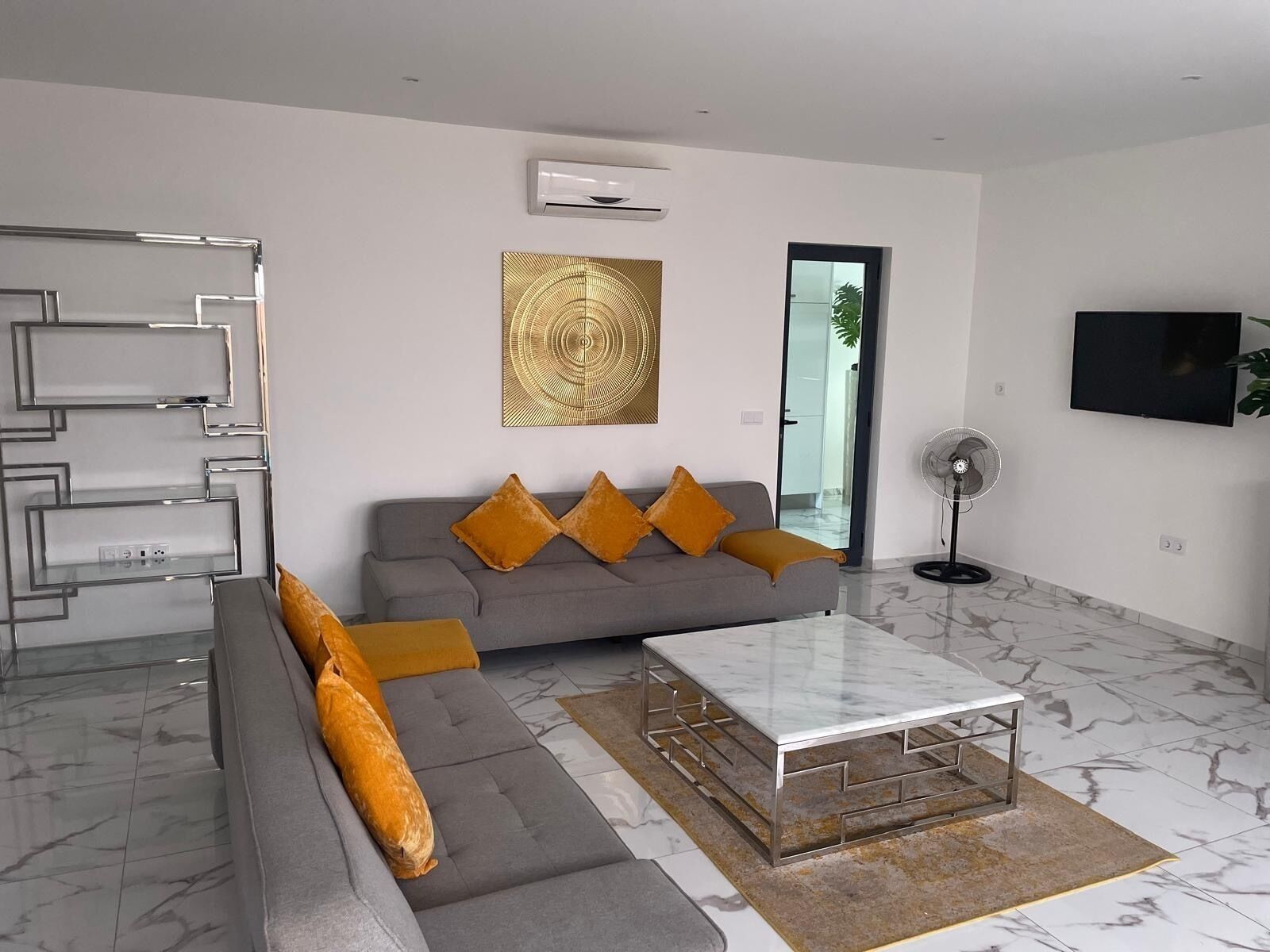 Een moderne woonkamer met een grijze bank, oranje kussens, een glazen salontafel, een vloerlamp en een aan de muur bevestigde televisie.