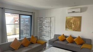 A modern living room with two grey sofas, each with four mustard yellow cushions, a glass coffee table, and a geometric wall art piece.