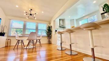 A modern kitchen with wooden flooring, a dining area with white chairs, a ceiling light fixture, and a window with blinds.