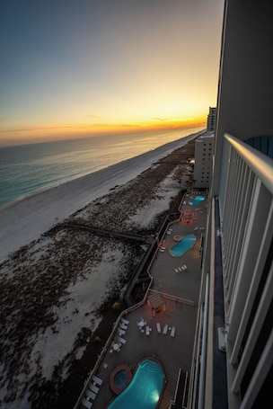 A balcony with a pool, lounge chairs, and a view of the beach at sunset.