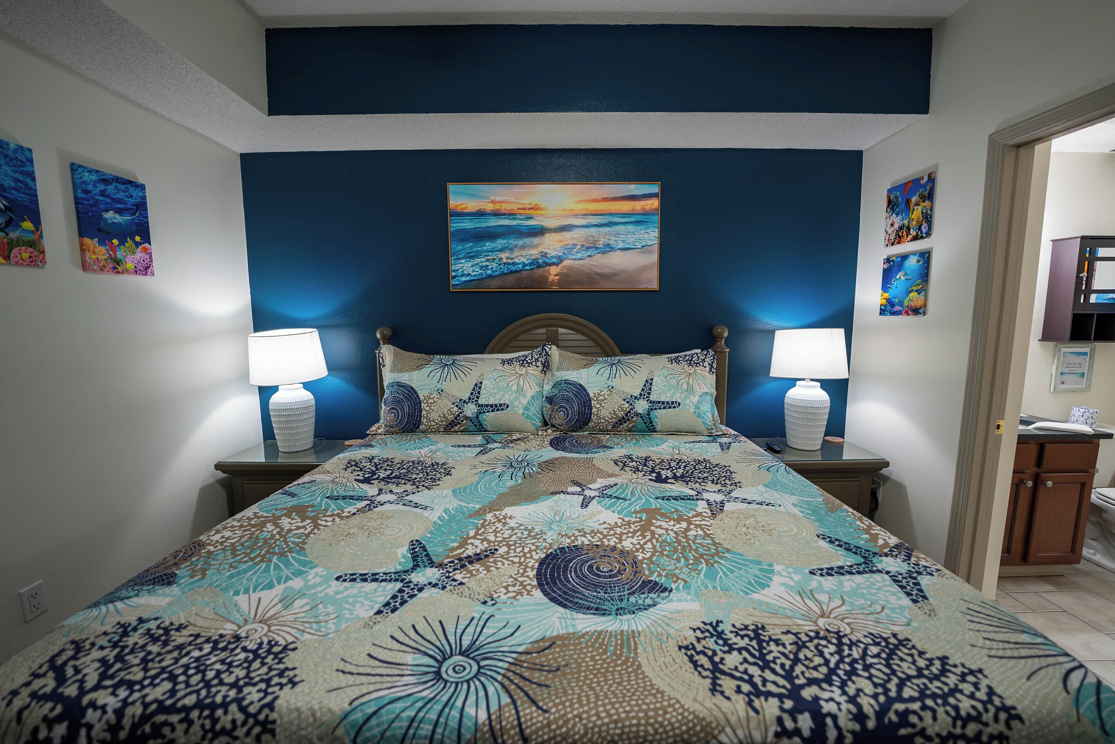 Une chambre à coucher avec un lit, deux lampes de chevet et un tableau représentant un coucher de soleil sur l’océan, accroché au mur.