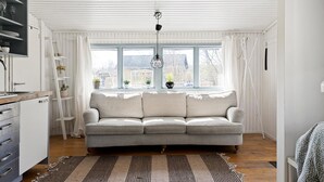 A spacious living room with a grey sofa, a wooden floor, a kitchen area with a sink and cabinets, and a large window with curtains. - OWN HOUSE, CLOSE TO WORK ESKILSTUNA STRÄNGNÄS, 1H STOCKHOLM, PARKING, WIFI, LAUNDRY (Södermanlands län)
