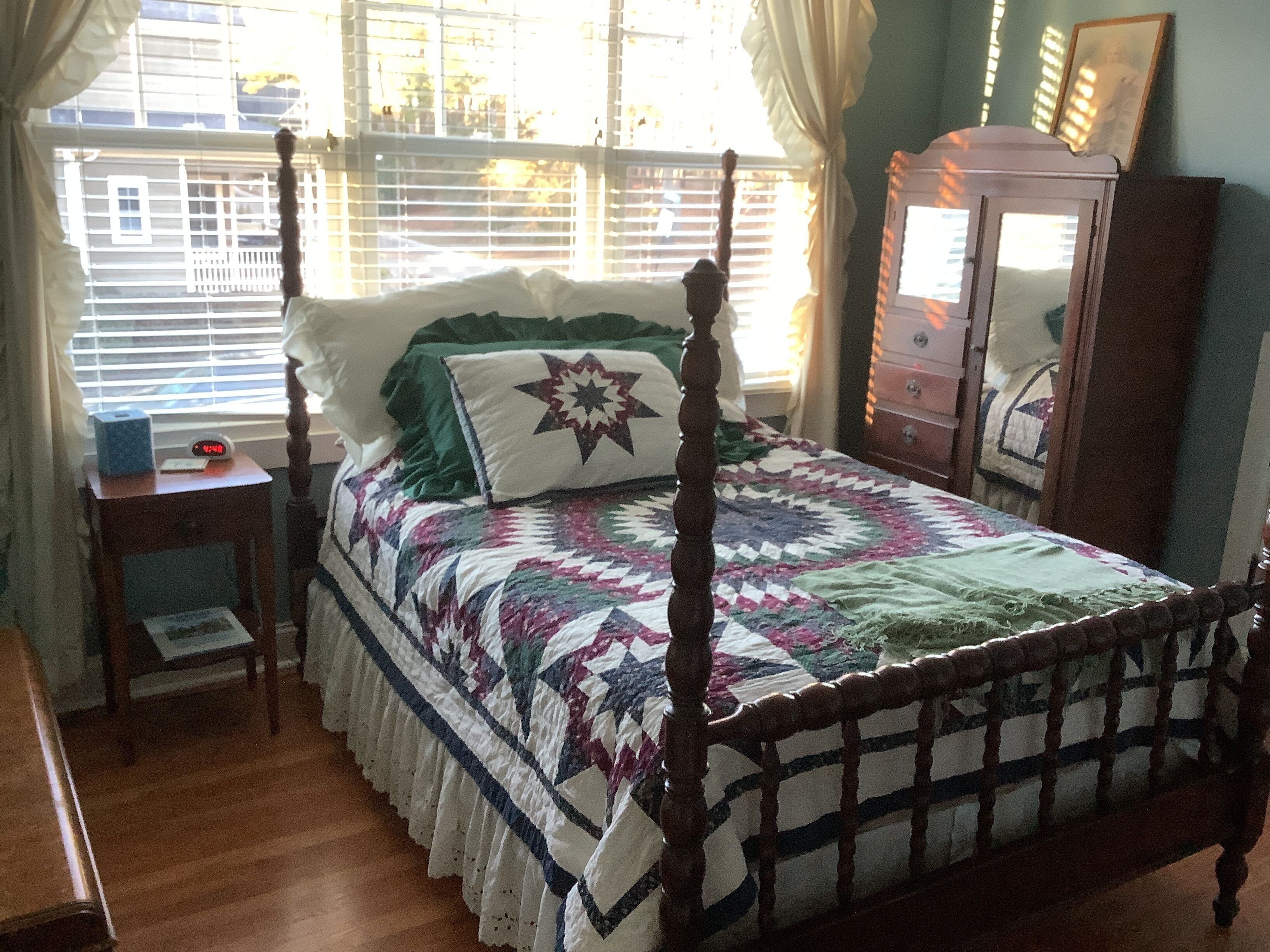A four-poster bed with a quilt, a bedside table with a clock, and a dresser with a mirror.