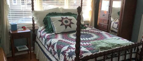 A four-poster bed with a quilt, a bedside table with a clock, and a dresser with a mirror.