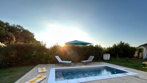 A swimming pool with two lounge chairs and a sun umbrella, surrounded by a wooden deck and greenery.