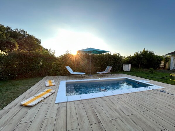 Une piscine avec deux chaises longues et un parasol, entourée d’une terrasse en bois et de verdure.