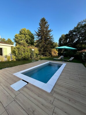 Une piscine rectangulaire avec une terrasse en bois, entourée de verdure et surplombée d’un ciel bleu clair.