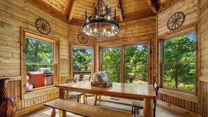 Ein Holzhütten-Interieur mit einem großen Esstisch, Bänken, einer Chandelüster und großen Fenstern mit Blick ins Freie.