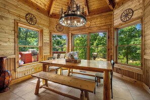 A wooden cabin interior with a large dining table, benches, a chandelier, and large windows offering a view of the outdoors.