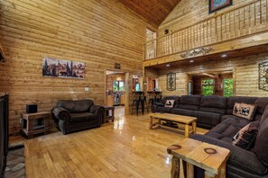 A log cabin interior with a wooden floor, two sofas, a coffee table, a TV, and a kitchen area visible through an open door.