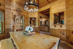 A wooden interior with a large wooden table, a chandelier, and a view of the outside through a window.