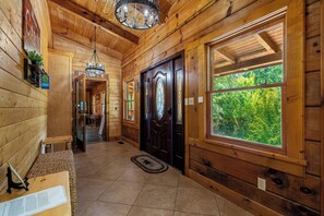 A wooden cabin interior with a bench, a door, a chandelier, and a view of greenery outside.