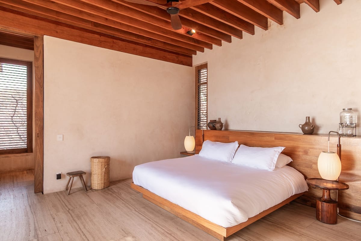 A spacious bedroom with a large bed, wooden ceiling beams, a small stool, and a bedside table with a lamp.