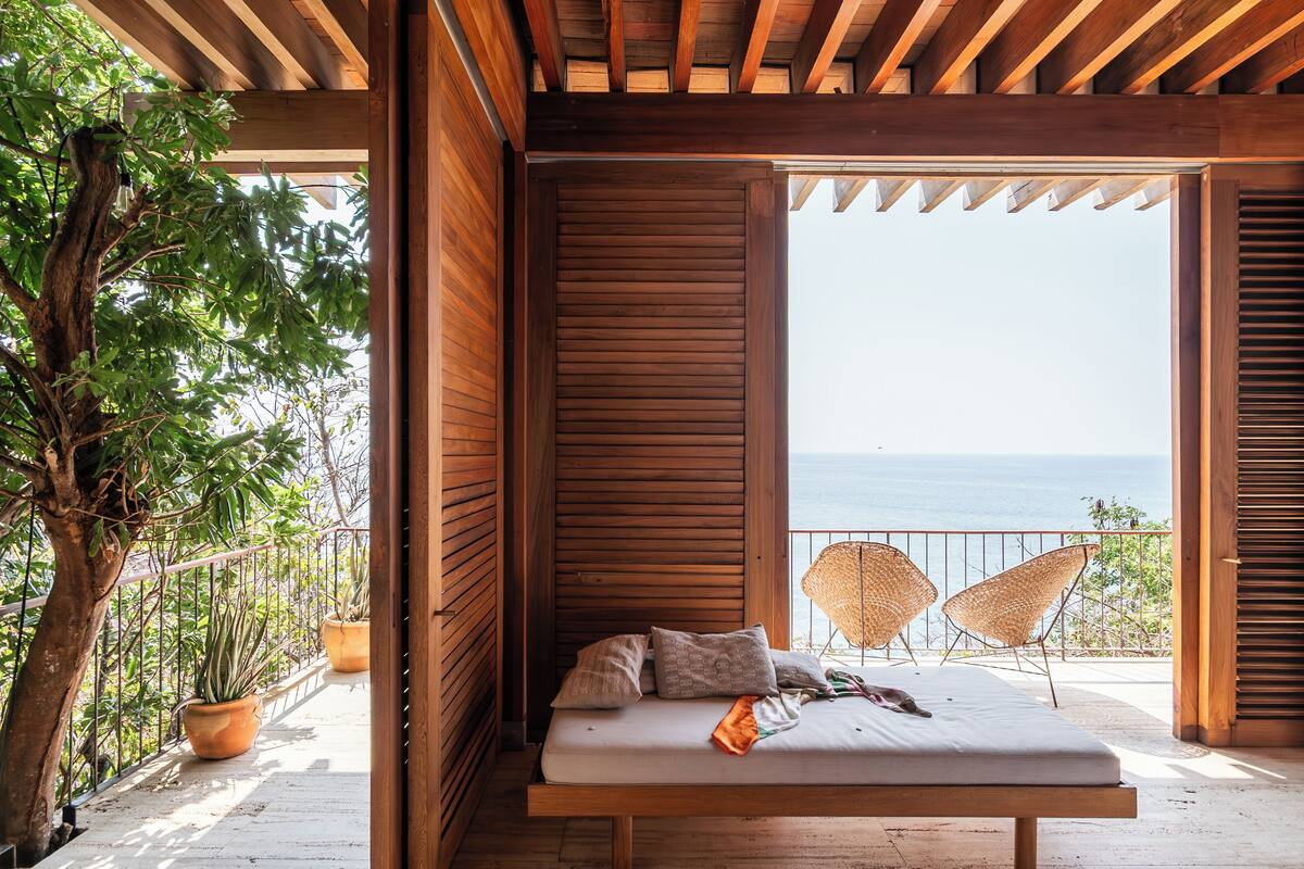 A wooden balcony with a bed, two wicker chairs, and a view of the sea.