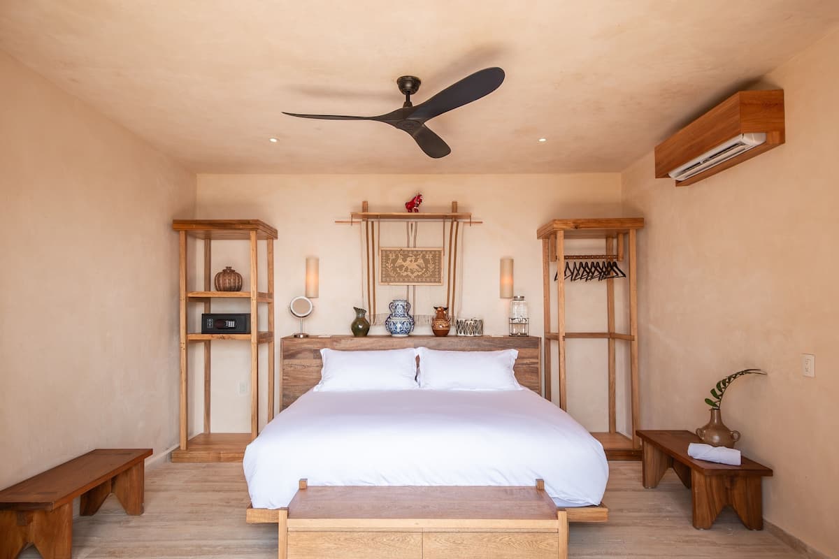 A bedroom with a large bed, wooden headboard, bedside tables, a ceiling fan, and a shelf with decorative items.