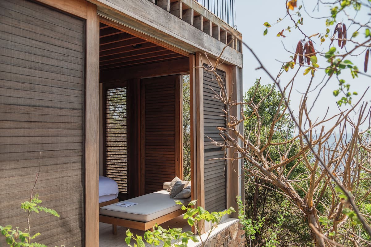 A wooden balcony with a bed, a window with blinds, and a view of trees and sky.