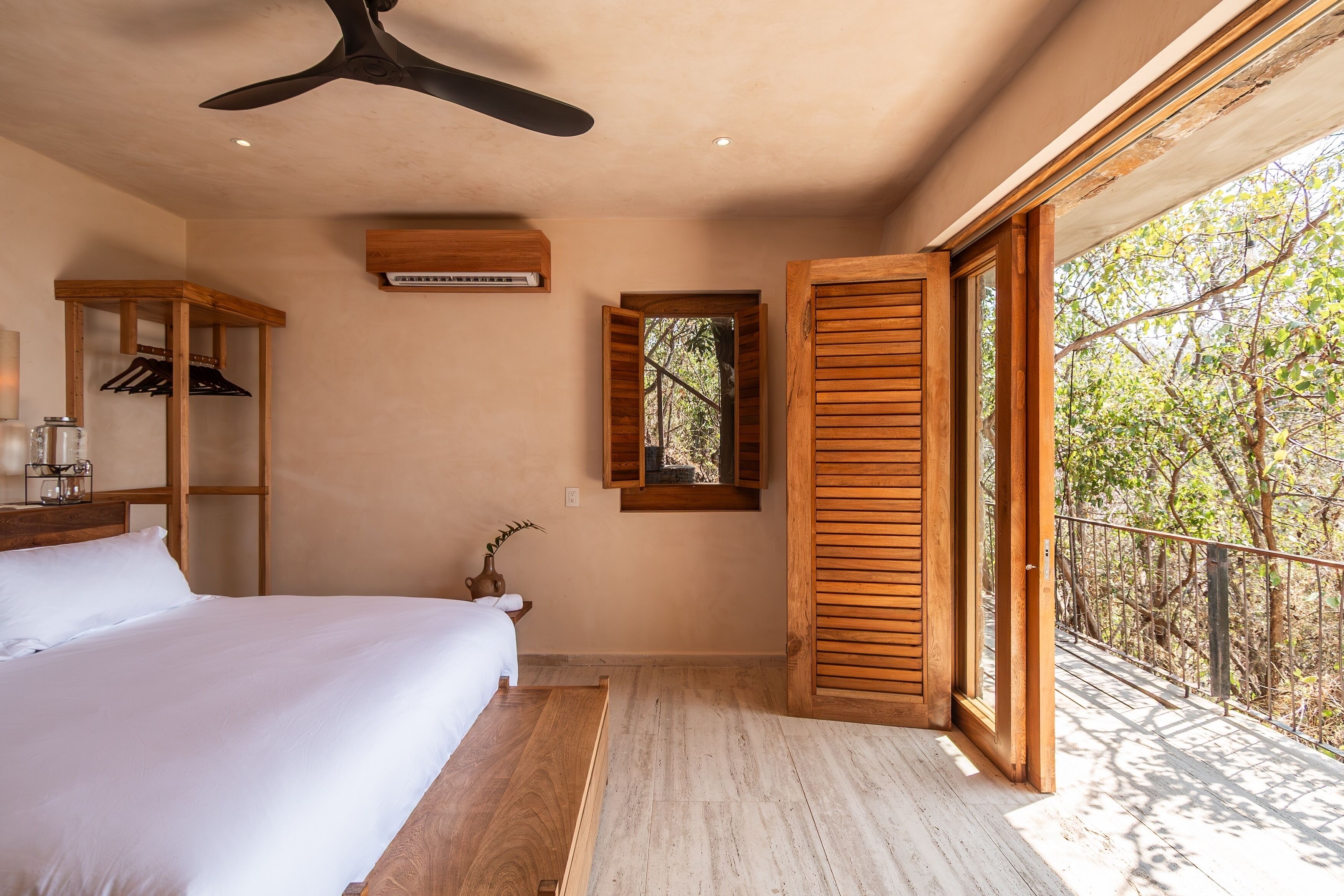 A bedroom with a bed, wooden headboard, a ceiling fan, a small window, and a balcony with a view of trees.