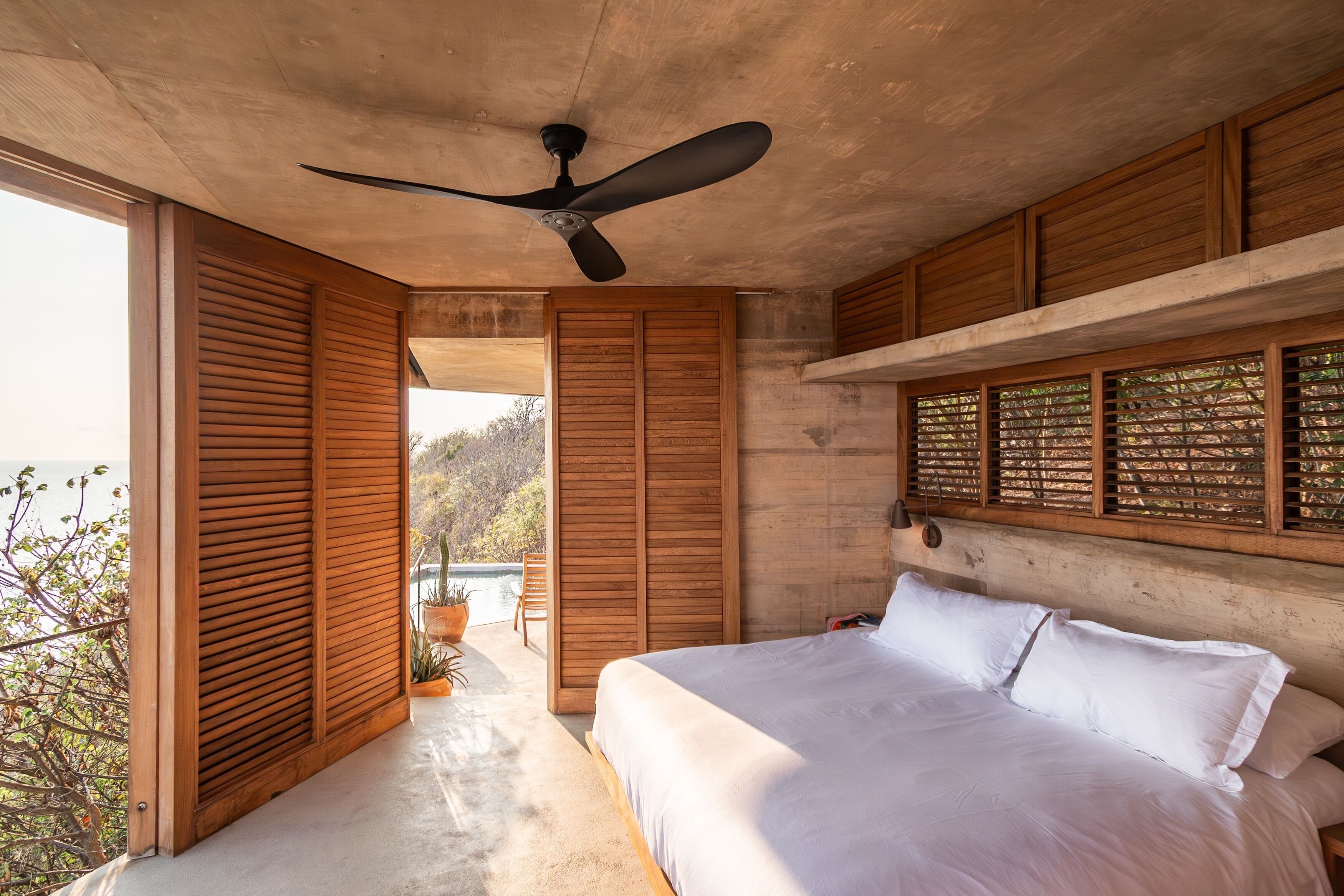 A bedroom with a large bed, wooden shutters, a ceiling fan, and a view of the outdoors.