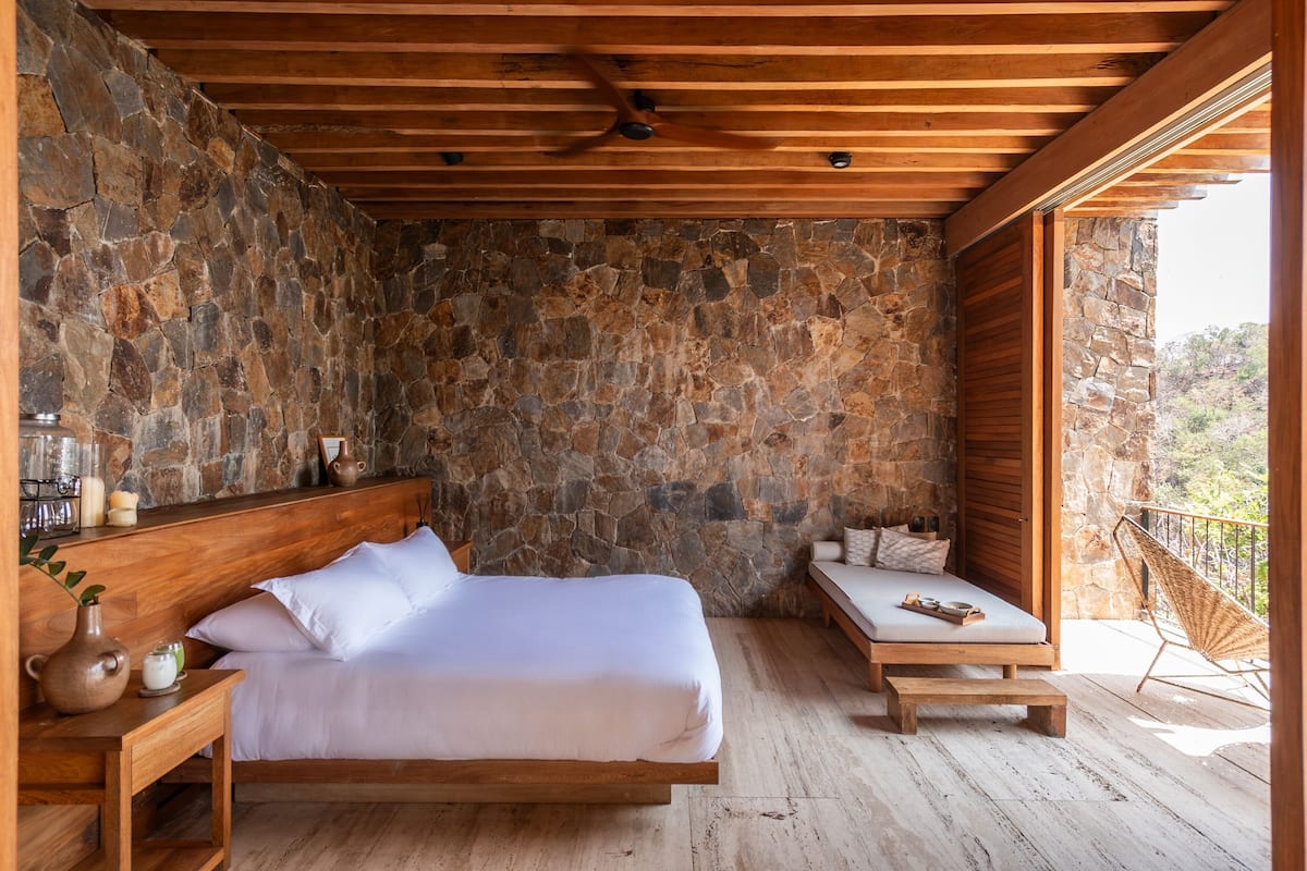 A bedroom with a stone wall, wooden ceiling, a bed, bedside table, and a balcony with a chair.