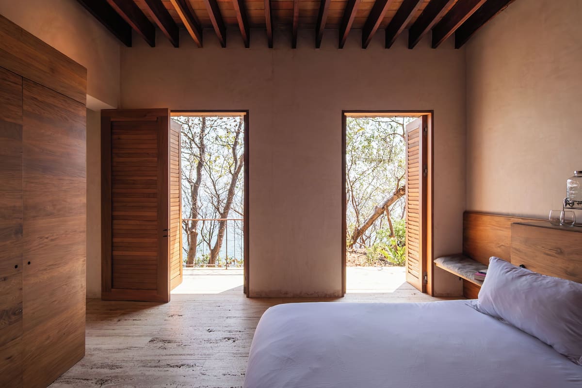 A bedroom with a wooden floor, a bed with white bedding, a wooden headboard, and a view of trees through open doors.