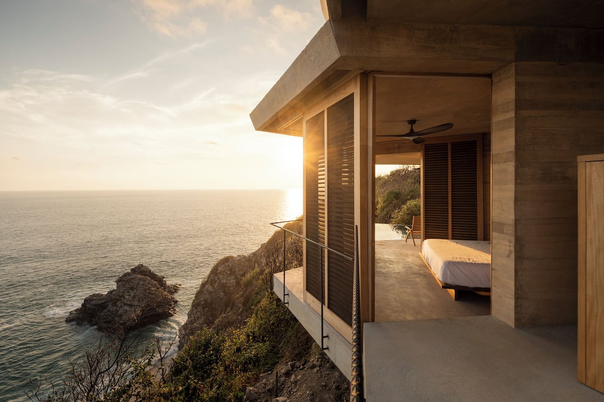 A modern bedroom with a large window overlooking the ocean, a ceiling fan, and a bed with a wooden headboard.