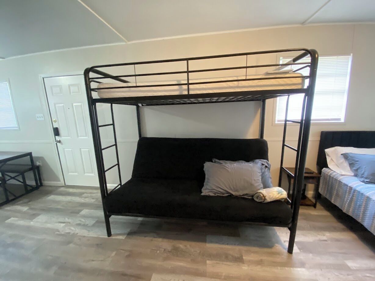 A bunk bed with a black frame, a black mattress, and a single pillow. There is a small table and a chair to the left, and a bed with a blue and white striped bedspread to the right.