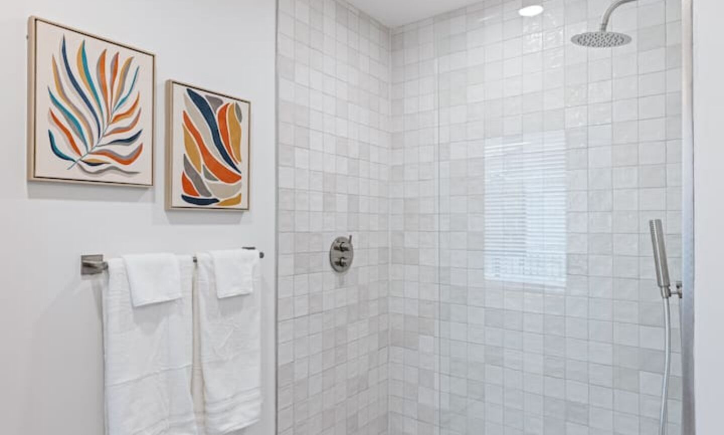 A modern bathroom with white tiles, a shower, and two abstract paintings on the wall.