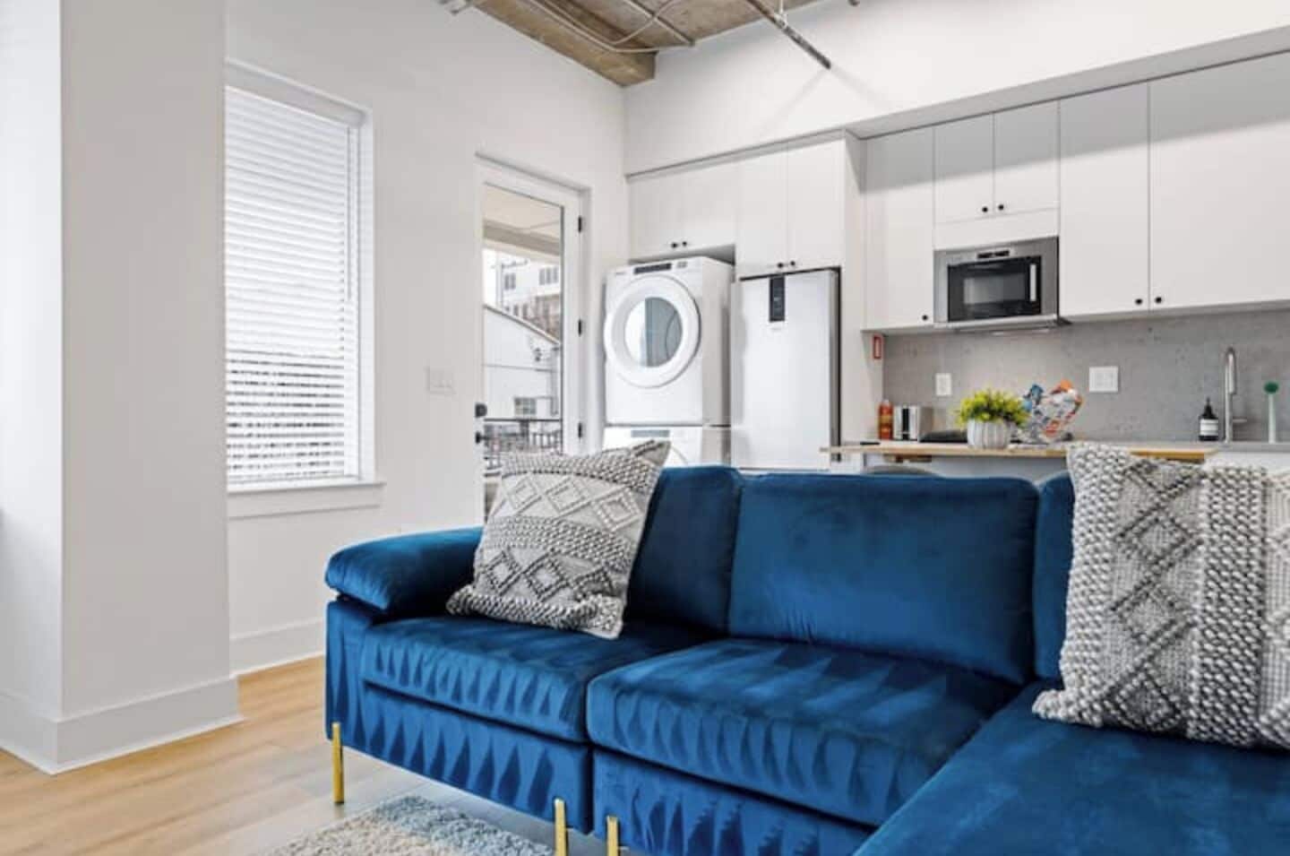 Un salon moderne avec un canapé bleu, un lave-linge et un coin cuisine doté de meubles et d’appareils électroménagers blancs.