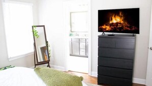 A bedroom with a bed, a dresser, a TV on a stand, and a door with a view of a kitchen.