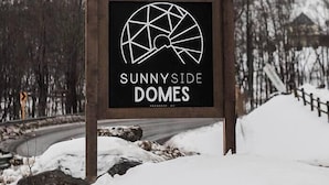 Opastekyltti, jossa lukee ”SUNNYSIDE DOMES”, ja jossa on geometrinen logo. Taustalla lumipeitteinen maisema, jossa on puita ja tie.