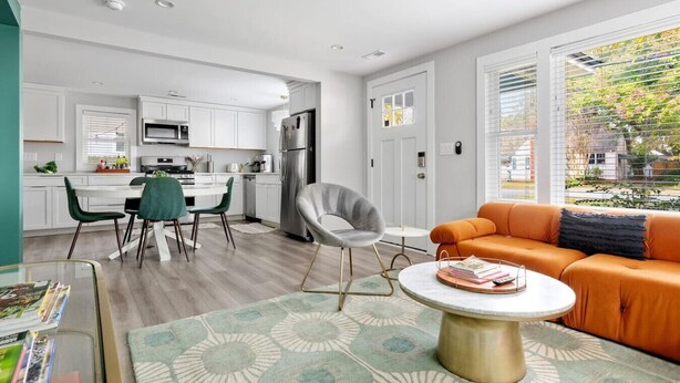 A modern kitchen with white cabinets, a dining area with a table and chairs, and a living room with an orange sofa and a coffee table.