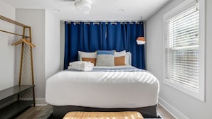 A neatly arranged bedroom with a large bed, blue curtains, a ceiling fan, and a window with blinds.