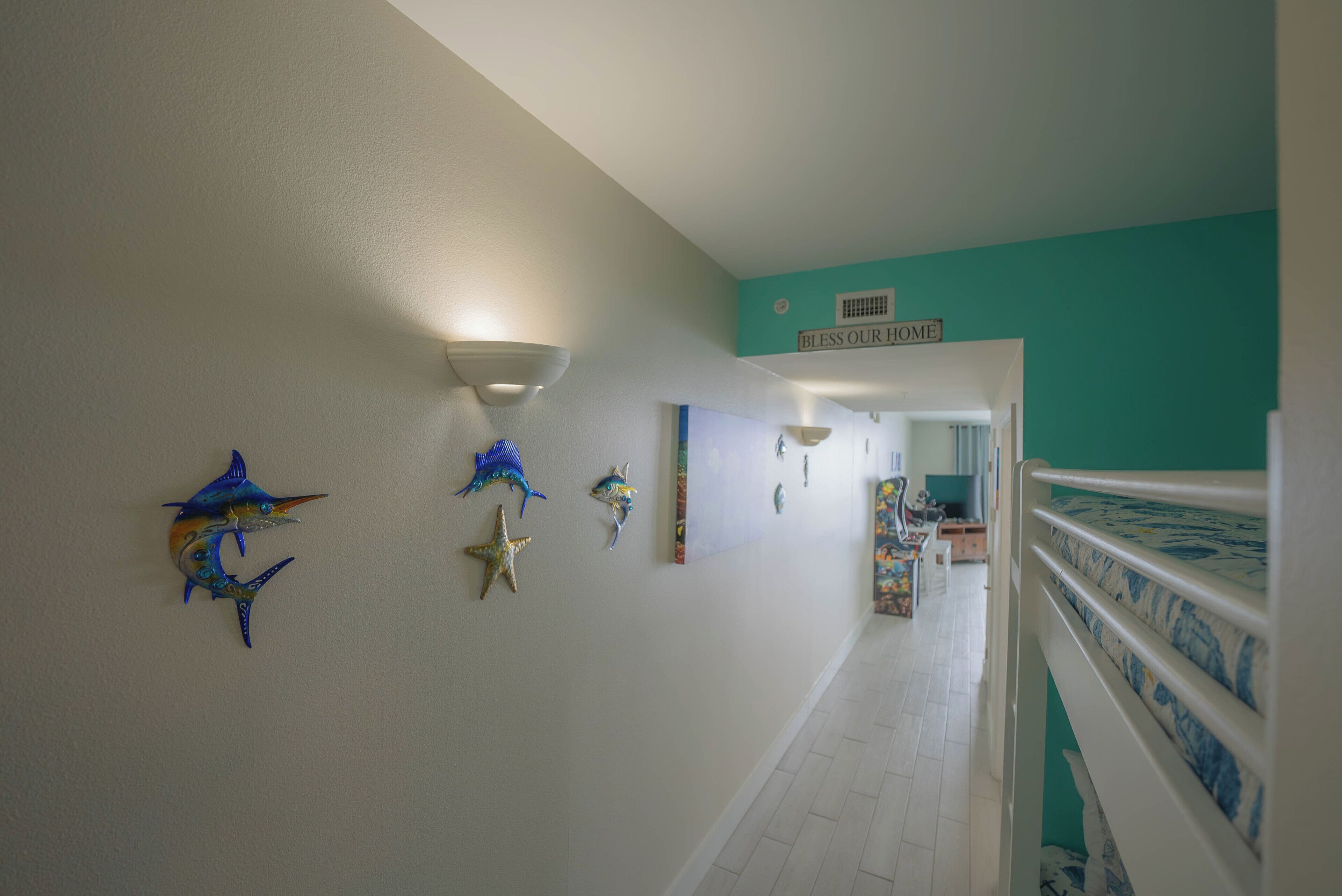 Un couloir agrémenté d’œuvres d’art murales, une pancarte sur laquelle on peut lire « Que notre foyer soit béni », et une vue sur une chambre à coucher avec une couvre-lit à motifs.