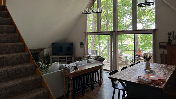 A spacious living area with a large window, a wooden staircase, a sofa, a dining table, and a television. - Charming lake front 3-bedroom/loft cabin w/ AC, WiFi in delightful Cub Run, KY. (Cub Run)