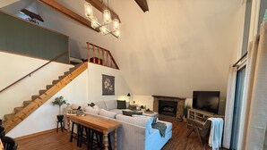A spacious living room with a wooden staircase, a fireplace, and a television. - Charming lake front 3-bedroom/loft cabin w/ AC, WiFi in delightful Cub Run, KY. (Cub Run)
