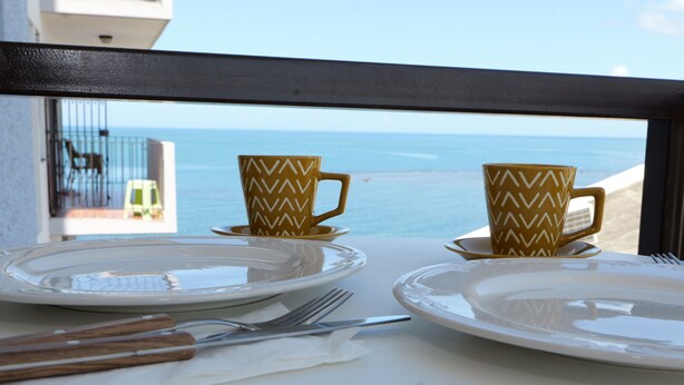 Taula preparada amb plats, coberts i gots estampats, amb vista a una platja i l'oceà.