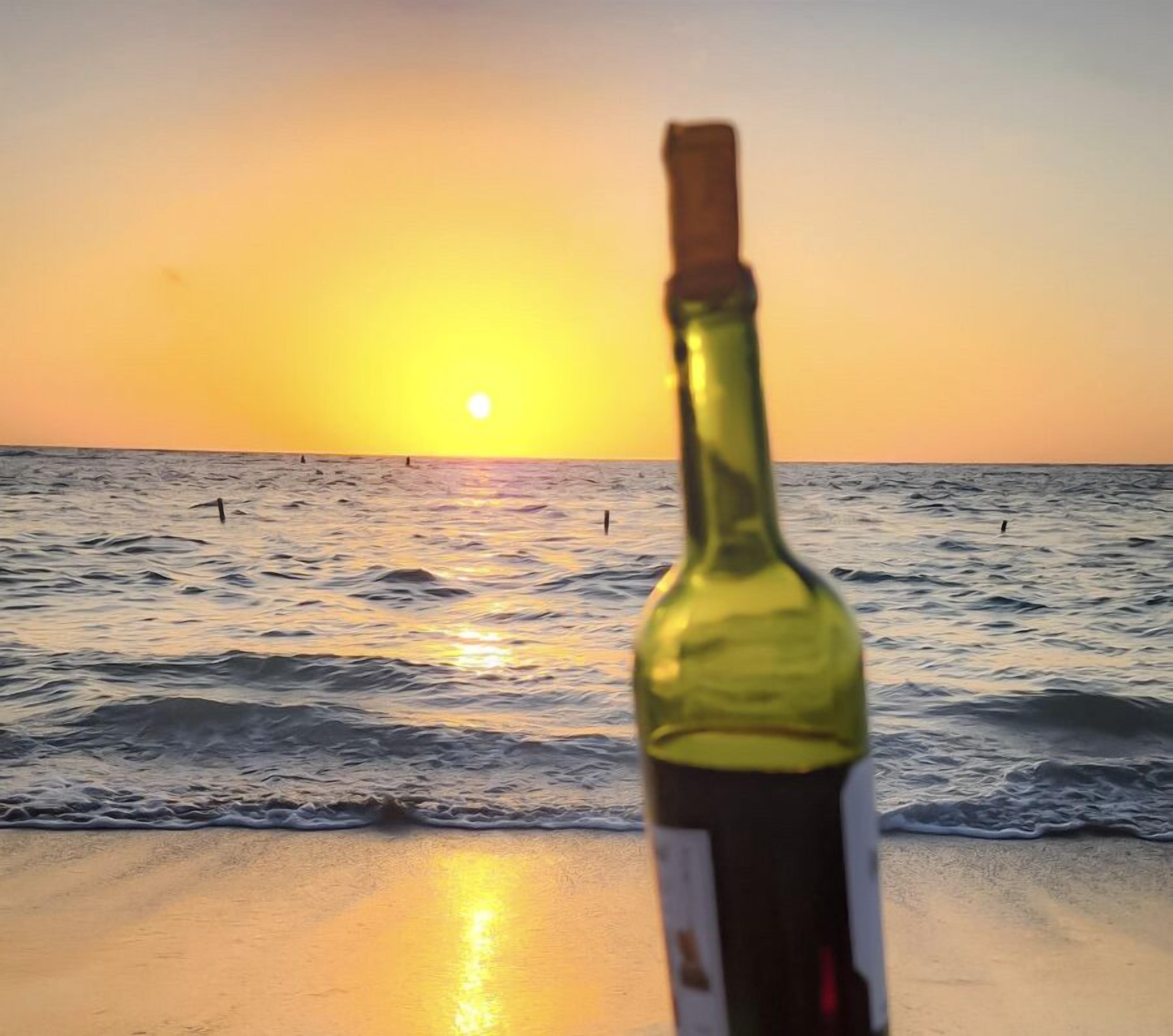 Una botella de vino en la playa con el océano y una puesta de sol al fondo.